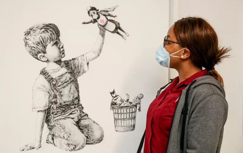 (FILES) A handout picture received from University Hospital Southampton on May 7, 2020 shows a member of staff posing with an artwork by street artist Banksy called "Game Changer", showing a boy playing with a nurse superhero toy with figures of Batman and Spiderman discarded in a basket as a tribute to NHS staff who are continuing to work during the COVID-19 pandemic, on a wall at the University Hospital Southampton, southern England. - painting by the British street artist Banksy sold for a record £16.75 million ($23.1 million, 19.4 million euros) on March 23, 2021, with proceeds going to the state-run health service. The painting, "Game Changer", sold for £14.4 million plus costs -- the highest ever for a Banksy painting -- after fierce competition between bidders in a nail-biting sale at Christie's auction house in London. (Photo by Stuart MARTIN / UNIVERSITY HOSPITAL SOUTHAMPTON / AFP) / RESTRICTED TO EDITORIAL USE - MANDATORY CREDIT "AFP PHOTO / UNIVERSITY HOSPITAL SOUTHAMPTON / STUART MARTIN " - NO MARKETING - NO ADVERTISING CAMPAIGNS - MANDATORY MENTION OF ARTIST UPON PUBLICATION - DISTRIBUTED AS A SERVICE TO CLIENTS