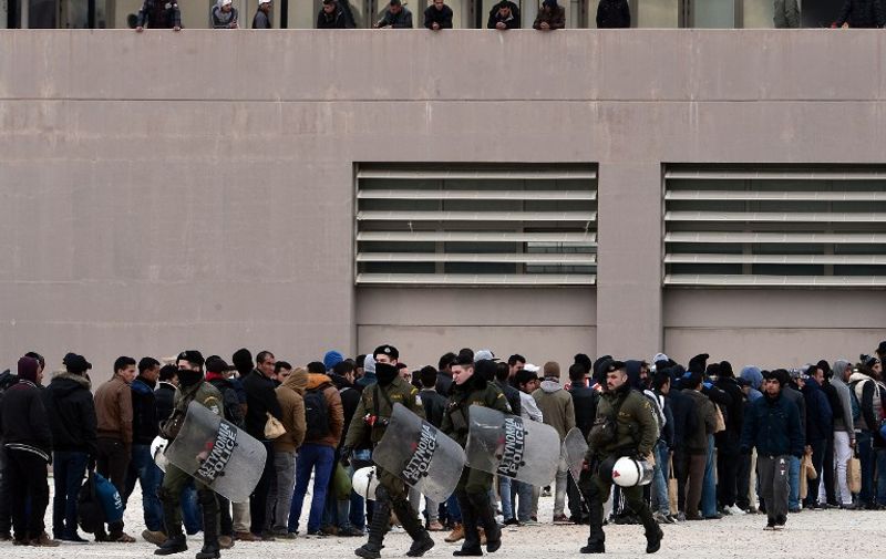 Riot police walks past migrants and refugees lining up outside a temporary housing facility for migrants located in a former Olympic hall in Faliro suburb of Athens on December 11, 2015. 
The venue houses mainly African and Iranian migrants, who were transfered earlier this week from Greece's borders with Macedonia, which allows only Syrians, Afghans and Iraqis through . All are trapped in Greece as economic migrants  by EU refugee rules,  and desperate to get out. Greece on Thursday said it would seek to deport economic migrants, who had been blocked at its border with Macedonia after Skopje clamped down on entry, if they are not entitled to asylum. / AFP / LOUISA GOULIAMAKI