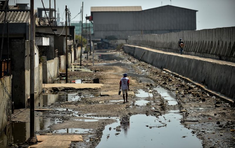 In this picture taken on July 12, 2019 a man walks on a muddy road between abandoned warehouses and a giant sea wall in northern Jakarta. Indonesia's capital is being swallowed into the ground at such an alarming rate that experts warn much of it could be submerged by 2050. (Photo by BAY ISMOYO / AFP) / TO GO WITH STORY: Indonesia-environment-social, by Dessy SAGITA