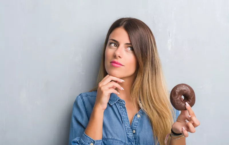 Young adult woman over grey grunge wall eating chocolate donut serious face thinking about question, very confused idea