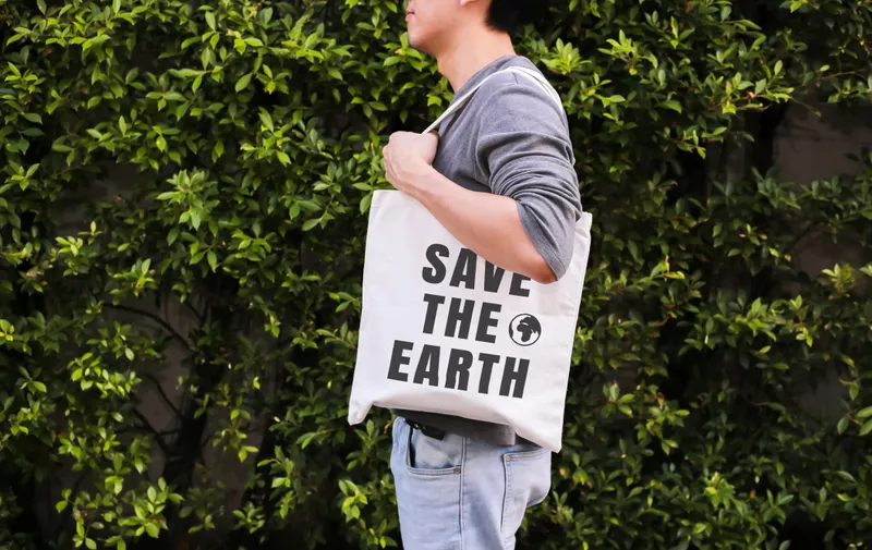 Young male hipster holding and carrying the Save The Earth tote handbag in green nature environment background - ecology and recycle concept