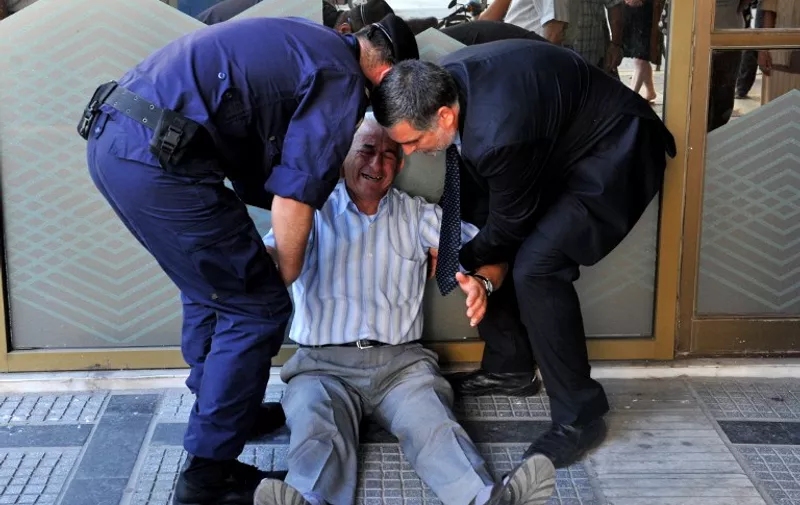 (FILES)-A July 3, 2015 shows Giorgos Chatzifotiadis, assisted by an employee and a policeman, as he sits on the ground outside a national bank branch, as pensioners (unseen) queue to withdraw their pensions, with a limit of 120 euros, in Thessaloniki.  Retiree Giorgos Chatzifotiadis had queued up at three banks in Greece's second city of Thessaloniki in the hope of withdrawing a pension on behalf of his wife, but all in vain.  When he was told at the fourth that he could not withdraw his 120 euros ($133), it was all too much and he collapsed in tears. AFP PHOTO /SAKIS MITROLIDIS