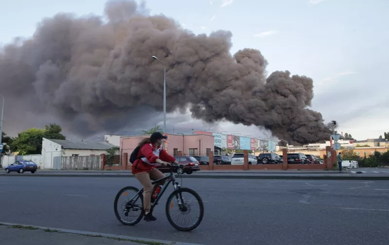 A woman riding a bicycle drives past a cloud of smoke from a fire in the background after a missile strike on a warehouse of an industrial and trading company in Odessa on July 16, 2022, amid the Russian invasion of Ukraine. (Photo by Oleksandr GIMANOV / AFP)