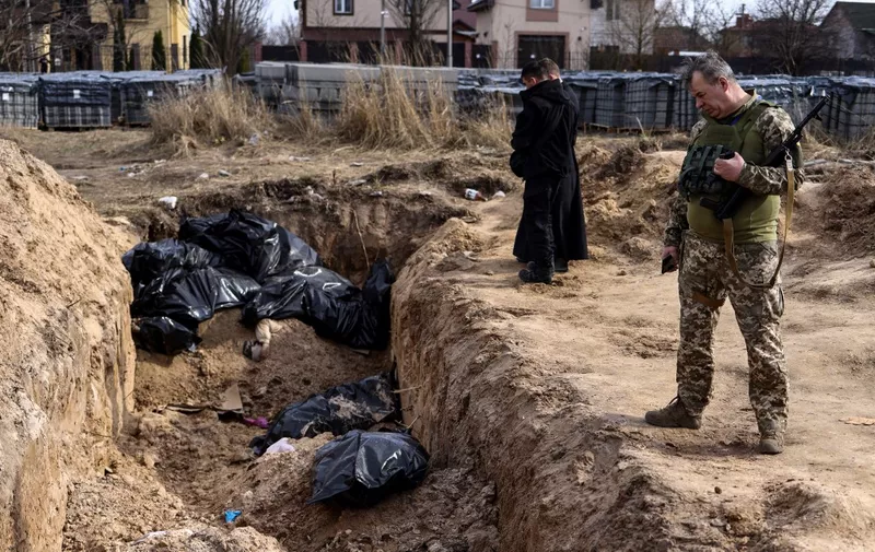 A Ukranian soldier looks at body bags as priests pray at a mass grave in the garden surrounding the St Andrew church in Bucha, on April 7, 2022, amid Russia's military invasion launched on Ukraine. - The UN humanitarian chief said on April 7 during a visit to the town of Bucha outside the Ukrainian capital Kyiv, which included a stop at the site of a mass grave that Ukrainians had dug near a church, that investigators would probe civilian deaths uncovered after Russian troops withdrew. (Photo by RONALDO SCHEMIDT / AFP)