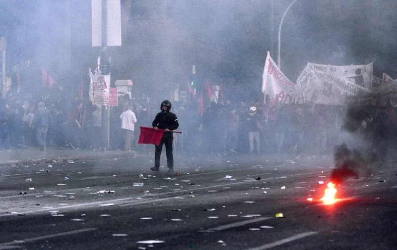 A protester stands with a flag amid stun grenades thrown by police during a protest rally against the latest reform measures demanded by Greece's creditors, in front of the Greek parliament building, in Athens on May 8, 2016.
The reforms to be voted on by MPs later on May 8 would reduce Greece's highest pension payouts, merge several pension funds, increase contributions and raise taxes for those on medium and high incomes. / AFP PHOTO / LOUISA GOULIAMAKI