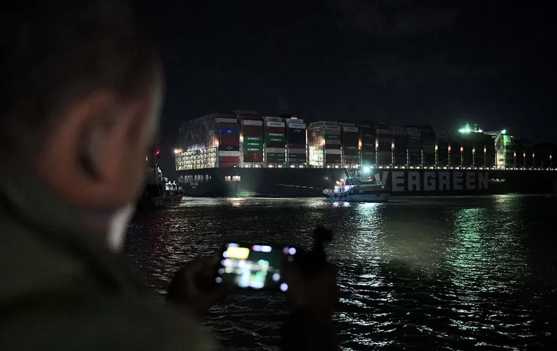 This picture taken late on March 27, 2021 shows a view of a man using his phone to take a picture of tugboats by the Panama-flagged MV 'Ever Given' (operated by Taiwan-based Evergreen Marine) container ship, which has been wedged diagonally across the span of the canal about six kilometres north of the Suez Canal's entrance by the Red Sea port city of Suez since March 23, blocking the waterway in both directions. (Photo by Ahmed HASAN / AFP)