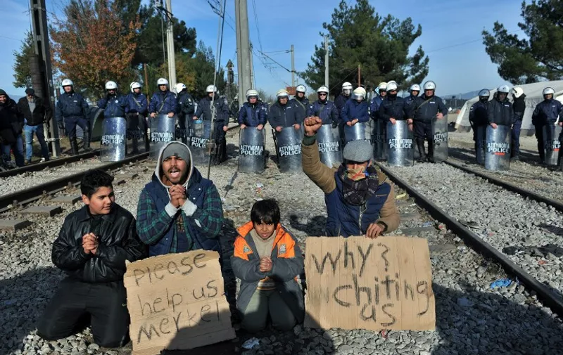 Greek police officers stand guard behind two men and two children demonstrating between railway tracks as they wait with other migrants and refugees to cross the Greek-Macedonian border near Idomeni on December 7, 2015.
Macedonia has restricted passage to northern Europe to only Syrians, Iraqis and Afghans who are considered war refugees. All other nationalities are deemed economic migrants and told to turn back, leaving over 1,500 people stuck on the border. / AFP / SAKIS MITROLIDIS