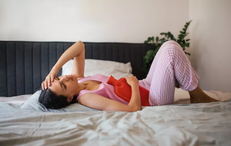Žena leži na krevetu s crvenom toplom vrećicom na trbuhu, držeći ruku na čelu zbog menstrualnih bolova