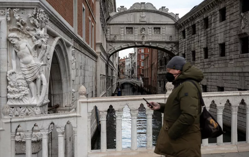 A man wearing a face mask and walks with his mobile phone as he walks past the bridge of Sighs on January 9, 2022 in Venice. (Photo by MARCO BERTORELLO / AFP)
