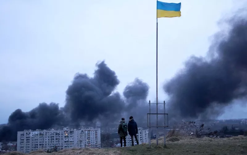 People stand in front of an Ukrainian national flag fluttering while watching dark smoke and flames rising from a fire following an air strike in the western Ukrainian city of Lviv, on March 26, 2022. - At least five people wounded in two strikes on Lviv, the regional governor said, in a rare attack on a city that has escaped serious fighting since Russian troops invaded last month. (Photo by Aleksey Filippov / AFP)