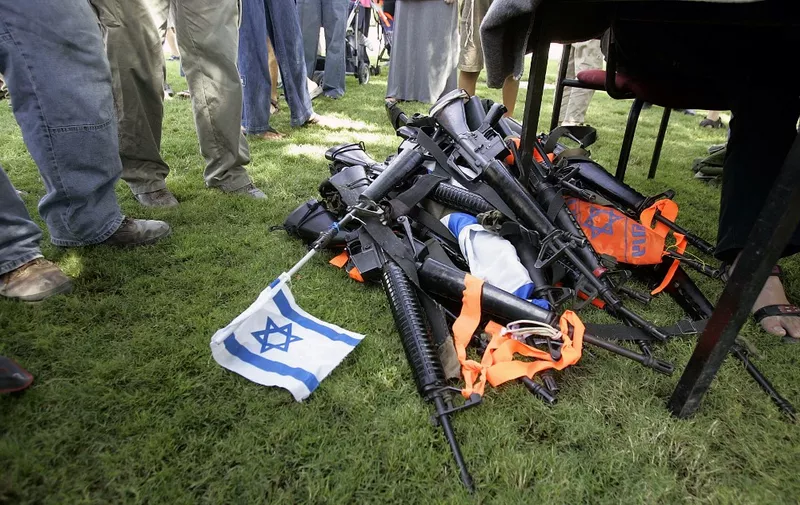 Israelis stand next to a pile of weapons turned in by resident settlers in the Southern Gaza Strip settlement of Ganei Tal 08 August 2005 in preparation for Israel's upcoming disengagement. The countdown is quickening for Jewish Israelis in Gaza to pack their bags and leave voluntarily during Israel's first-ever withdrawal from Palestinian territory. The operation to uproot the 8,000 Gaza settlers and residents of four small Israeli enclaves in the northern West Bank is due to begin on August 17.     AFP PHOTO/ Roberto SCHMIDT (Photo by Roberto SCHMIDT / AFP)