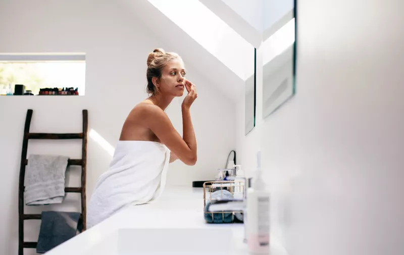 Young woman wrapped in towel looking in the mirror and applying cream on her face. Female taking care of her face skin in bathroom after bath.
