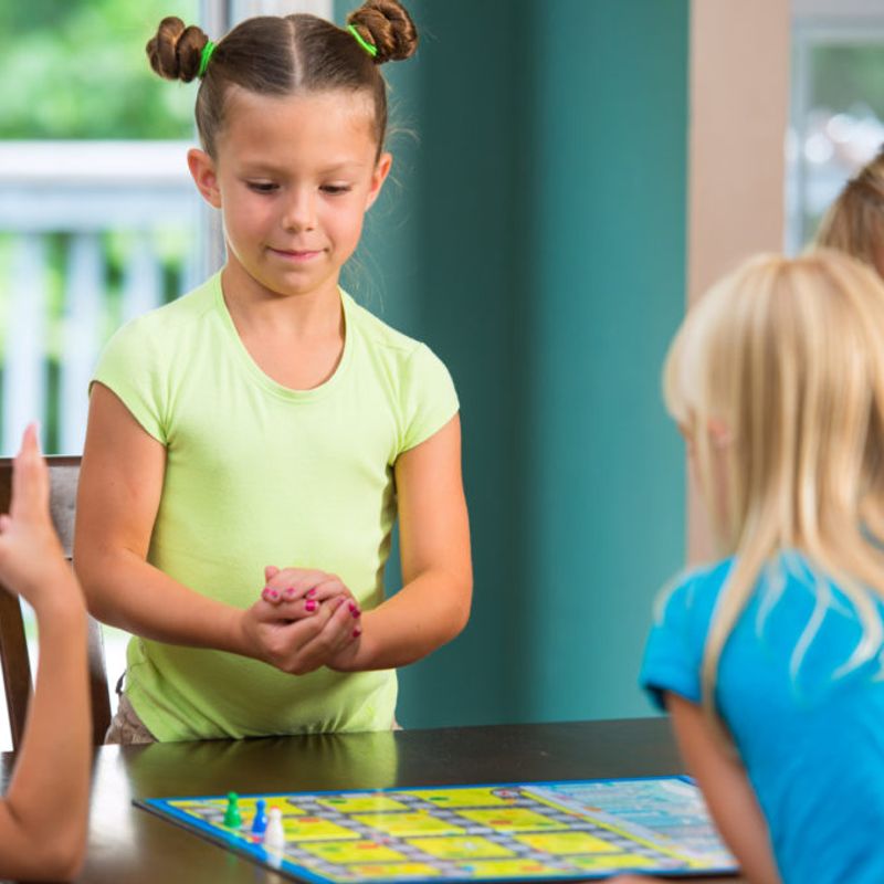 Authentic, fun image of a family playing a board game in a typical american home. Little girl with buns in hair shakes dice in her hands before her roll.