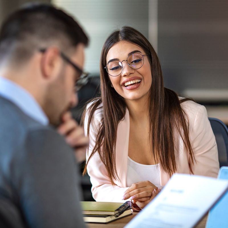 Portrait of young  female client or candidate sitting at table, talking to male manager and smiling in office. Job interview or consultancy concept. Young attractive woman during job interview