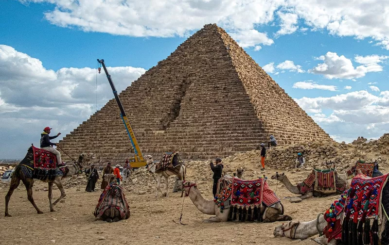 A crane lifting stones is pictured behind camels resting with their trainers by the Pyramid of Menkaure (or Menkheres, built in the 26th century BC) at the Giza Pyramids Necropolis, west of Cairo, on January 29, 2024. In a video posted on Facebook on January 26, Mostafa Waziri, the head of Egypt's Supreme Council of Antiquities, showed workers setting blocks of granite on the base of the pyramid, dubbing it "the project of the century." When originally built, the pyramid was encased in granite, but over time lost part of its covering. The renovation aims to restore the structure's original style by reconstructing the granite layer. (Photo by Khaled DESOUKI / AFP) / The erroneous mention[s] appearing in the metadata of this photo by Khaled DESOUKI has been modified in AFP systems in the following manner: [2024] instead of [2023]. Please immediately remove the erroneous mention[s] from all your online services and delete it (them) from your servers. If you have been authorized by AFP to distribute it (them) to third parties, please ensure that the same actions are carried out by them. Failure to promptly comply with these instructions will entail liability on your part for any continued or post notification usage. Therefore we thank you very much for all your attention and prompt action. We are sorry for the inconvenience this notification may cause and remain at your disposal for any further information you may require.