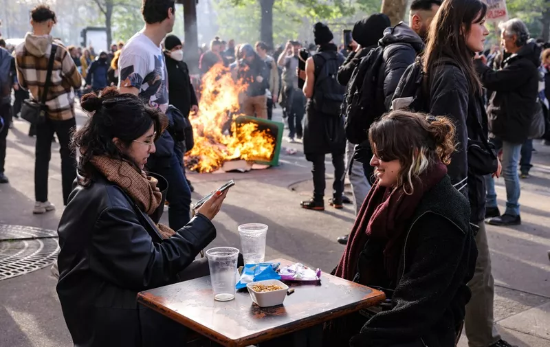 Two girls have a drink at a cafe terrace as a garbage can is set ablaze nearby during a demonstration on the 11th day of action after the government pushed a pensions reform through parliament without a vote, using the article 49.3 of the constitution, in Paris on April 6, 2023. - France on April 6, 2023 braced for another day of protests and strikes to denounce French President's pension reform one day after talks between the government and unions ended in deadlock. (Photo by Thomas SAMSON / AFP)