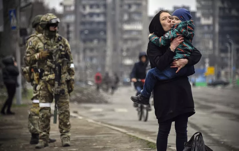 A woman holds and kisses a child next to Russian soldiers in a street of Mariupol on April 12, 2022, as Russian troops intensify a campaign to take the strategic port city, part of an anticipated massive onslaught across eastern Ukraine, while Russia's President makes a defiant case for the war on Russia's neighbour. - *EDITOR'S NOTE: This picture was taken during a trip organized by the Russian military.* (Photo by Alexander NEMENOV / AFP)