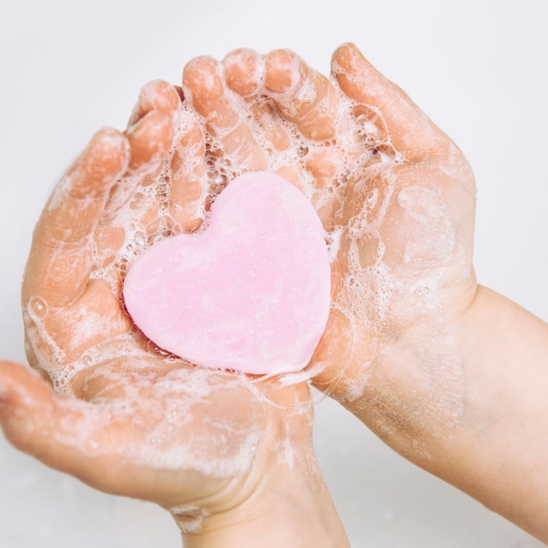 Importance of personal hygiene care. Flat lay view of child washing dirty hands with pink heart shape soap bar, lot of foam. Copy space.