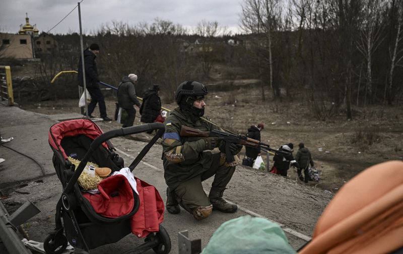 An Ukrainian serviceman takes cover as people evacuate  the city of Irpin on March 13, 2022. - Russian forces advance ever closer to the capital from the north, west and northeast. Russian strikes also destroy an airport in the town of Vasylkiv, south of Kyiv. A US journalist was shot dead and another wounded in Irpin, a frontline northwest suburb of Kyiv, medics and witnesses told AFP. (Photo by Aris Messinis / AFP)