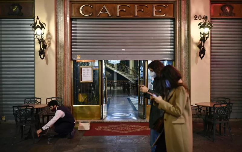 Two women walk past the "Caffe Torino" as a waiter prepares to close in downtown Turin, on October 26, 2020, as the country faces a second wave of infections to the Covid-19 (the novel coronavirus). - Italy's Prime Minister Giuseppe Conte tightened nationwide coronavirus restrictions on October 25, 2020 after the country registered a record number of new cases, despite opposition from regional heads and street protests over curfews. Cinemas, theatres, gyms and swimming pools must all close under the new rules, which come into force on October 26, 2020 and run until November 24, while restaurants and bars will stop serving at 6pm, the prime minister's office said. (Photo by Marco BERTORELLO / AFP)