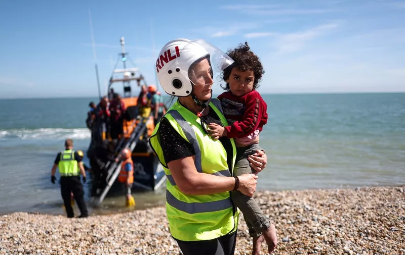 A member of the Royal National Lifeboat Institution carries a child as migrants, picked up at sea attempting to cross the English Channel from France, are helped ashore from an Royal National Lifeboat Institution (RNLI) lifeboat on the beach at Dungeness on the southeast coast of England on August 16, 2023. More than 100,000 migrants have crossed the Channel on small boats from France to southeast England since Britain began publicly recording the arrivals in 2018, official figures revealed on August 11, 2023. (Photo by HENRY NICHOLLS / AFP)