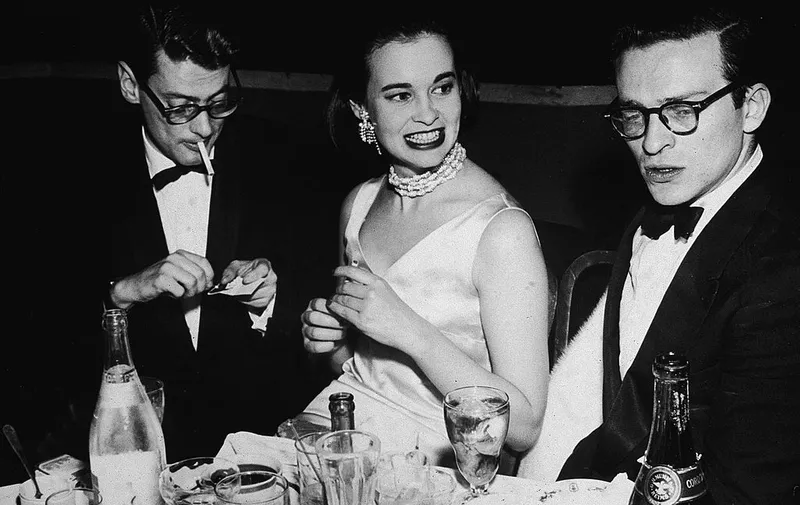 American photographer Richard Avedon (left, lighting a cigarette), American heiress and designer Gloria Vanderbilt, and American film director Sidney Lumet, sit at a table covered with glasses and bottles during a party for the premiere of the movie 'East of Eden' directed by Elia Kazan, 1955. (Photo by Getty Images)