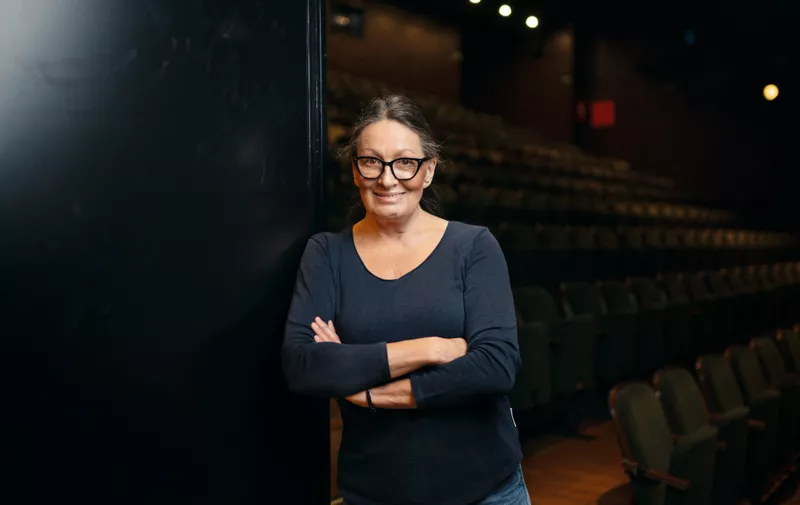 Žena srednje dobi s naočalama i tamnoplavom bluzom stoji prekriženih ruku ispred praznog kazališta.