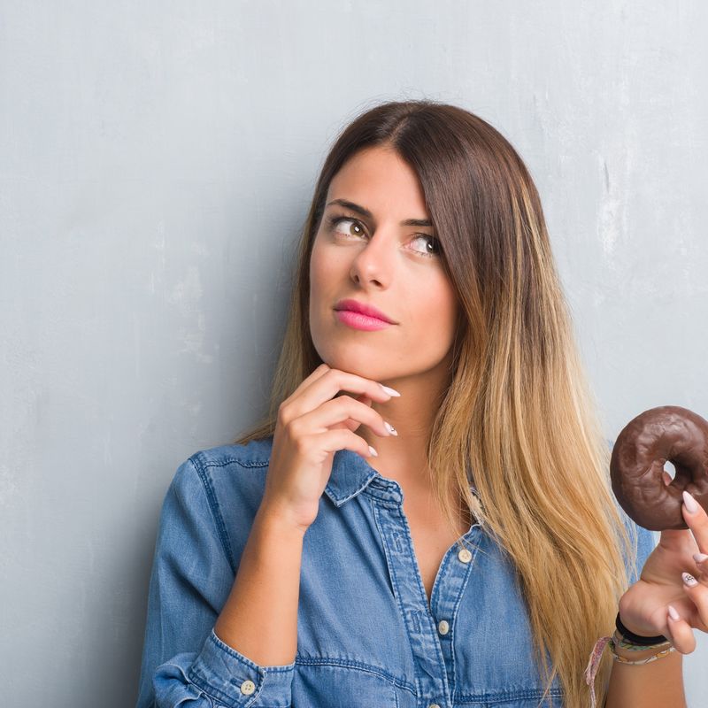 Young adult woman over grey grunge wall eating chocolate donut serious face thinking about question, very confused idea