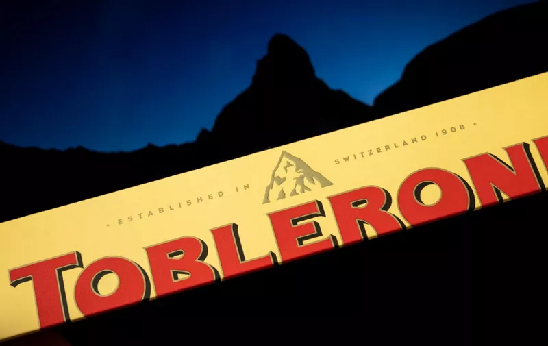 A picture taken on March 6, 2023 in Geneva shows a the packaging of a Toblerone chocolate bar owned by US firm Mondelez with a picture of the Matterhorn mountain seen in silhouette in the background. - Toblerone is to remove the Swiss iconic Matterhorn peak from its packaging when some of the chocolate's production is moved from Switzerland to Slovakia and replaced by a more generic mountain under strict "Swissness" rules. (Photo by Fabrice COFFRINI / AFP)