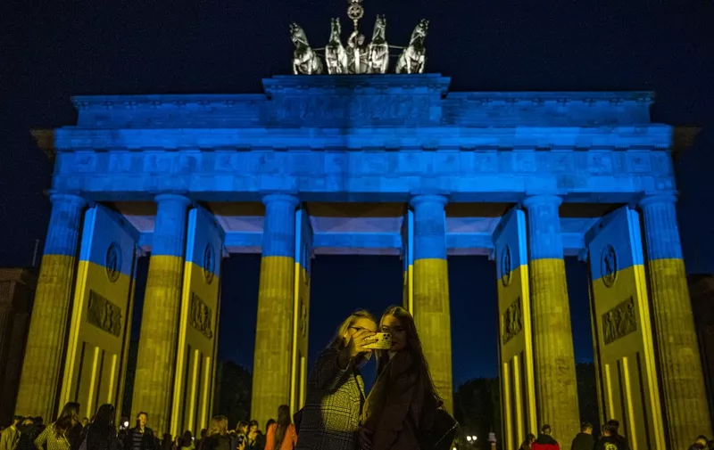 People take selfies in front of the landmark Brandenburg Gate illuminated in the colors of the Ukrainian flag in Berlin on May 9, 2022, to show solidarity with Ukraine amid the ongoing Russian invasion. (Photo by John MACDOUGALL / AFP)