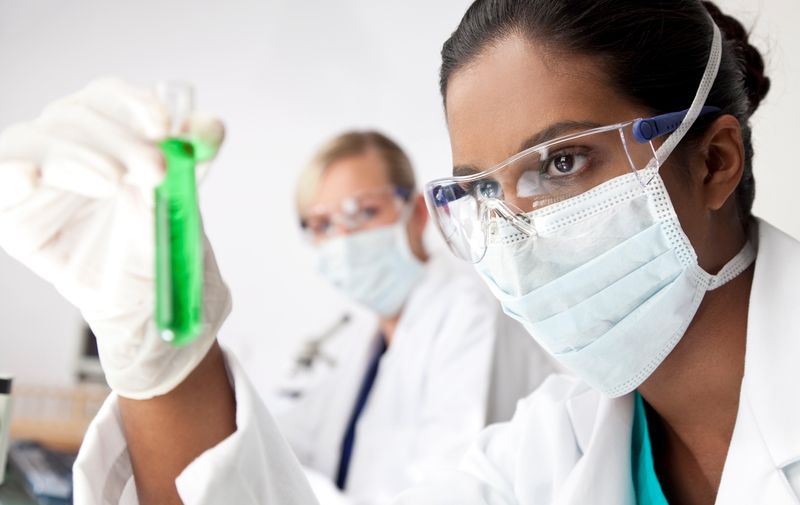 A female Asian medical or scientific researcher or doctor looking at a test tube of a green solution in a laboratory with her blond female colleague out of focus behind her.