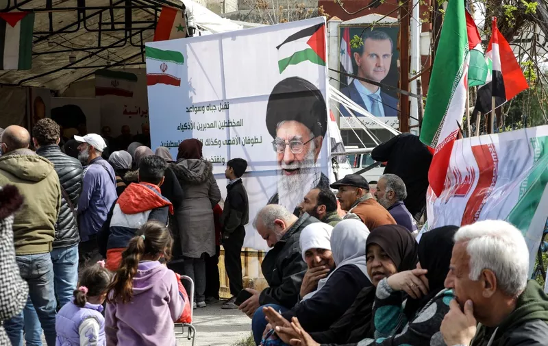 Portraits of Syria's President Bashar al-Assad (R) and Iran's supreme leader Ayatollah Ali Khamenei stand as Palestinians sit at the entrance of the Yarmuk camp for Palestinian refugees, south of Damascus, during a delivery of humanitarian aid provided by Iran as part of the Muslim holy fasting month of Ramadan on March 26, 2024. (Photo by LOUAI BESHARA / AFP)