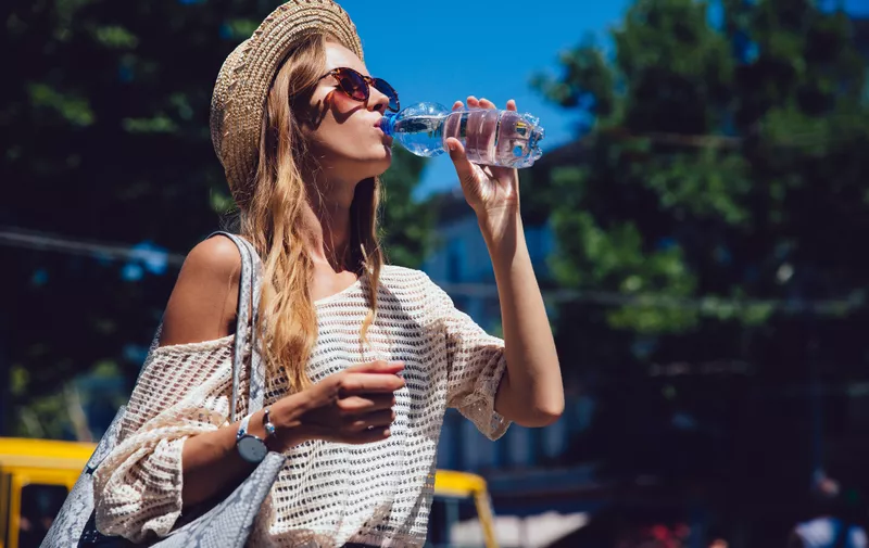 Profile of lovely woman in sunglasses, drinking a water, during walking outdoors. Dressed in trendy clothes, in hat. Summertime.
