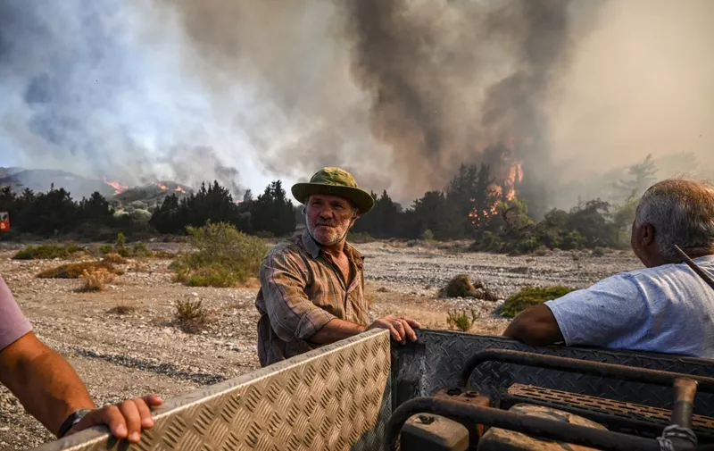 Men stand by their pick-up truck as they watch wildfires close to the village of Vati on the Greek island of Rhodes on July 25, 2023. Some 30,000 people fled the flames on Rhodes at the weekend, the country's largest-ever wildfire evacuation as the prime minister warned that the heat-battered nation was "at war" with several wildfires and spoke of three difficult days ahead. (Photo by Spyros BAKALIS / AFP)