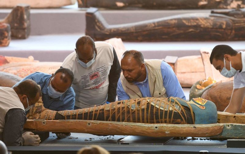 Archaeologists inspect a mummy, wrapped in a burial shroud adorned with brightly coloured hieroglyphic pictorials, during the unveiling of an ancient treasure trove of more than a 100 intact sarcophagi, at the Saqqara necropolis 30 kms south of the Egyptian capital Cairo, on November 14, 2020. - Egypt announced the discovery of an ancient treasure trove of more than a 100 intact sarcophagi, the largest such find this year. The sealed wooden coffins, unveiled on site amid fanfare, belonged to top officials of the Late Period and the Ptolemaic period of ancient Egypt. They were found in three burial shafts at depths of 12 metres (40 feet) in the sweeping Saqqara necropolis south of Cairo. (Photo by Ahmed HASAN / AFP)
