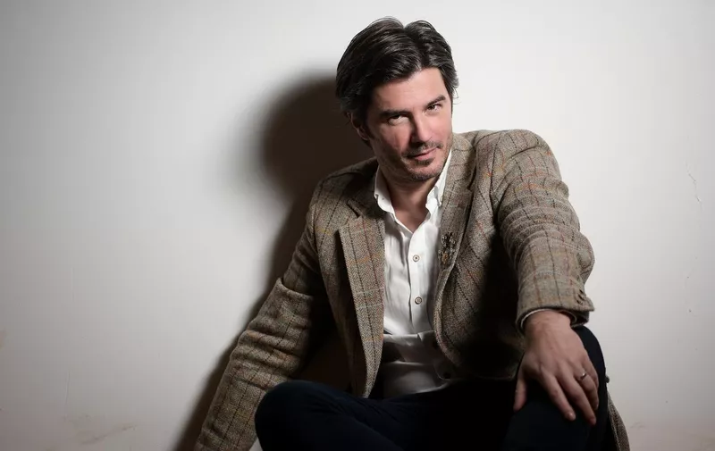 Muškarac tamne kose u smeđem karirastom sakou i bijeloj košulji sjedi opušteno na stolici uz bijelu pozadinu.