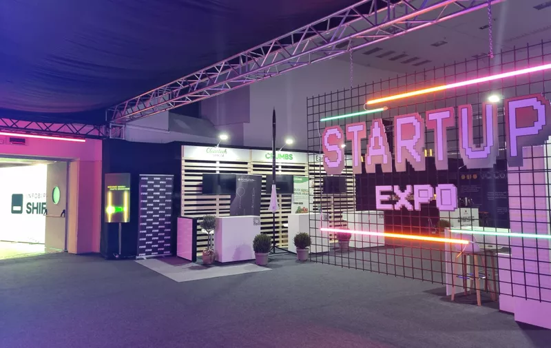 Moderno osvjetljena izložbena dvorana s ružičastim neonskim natpisom "STARTUP EXPO" na metalnoj rešetki.