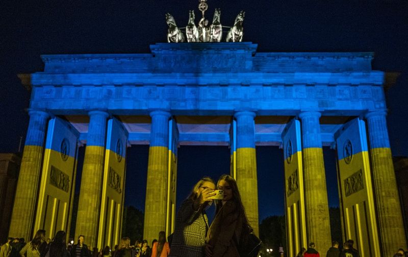 People take selfies in front of the landmark Brandenburg Gate illuminated in the colors of the Ukrainian flag in Berlin on May 9, 2022, to show solidarity with Ukraine amid the ongoing Russian invasion. (Photo by John MACDOUGALL / AFP)