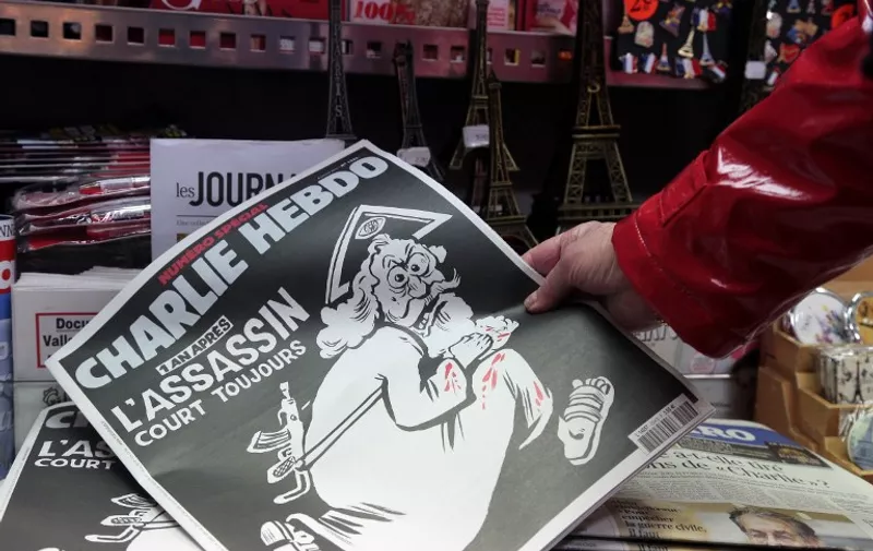 A woman looks at the special commemorative edition of French satirical newspaper Charlie Hebdo at a newsstand in Paris, on January 6, 2015, to mark the one-year anniversary of the jihadist attack that claimed the lives of 12 people, including three of its best-known cartoonists.
True to form, the cover is unabashedly provocative, featuring a Kalashnikov-toting God figure wearing a blood-stained white robe, under the headline: "One year on: The killer is still at large." / AFP / JACQUES DEMARTHON