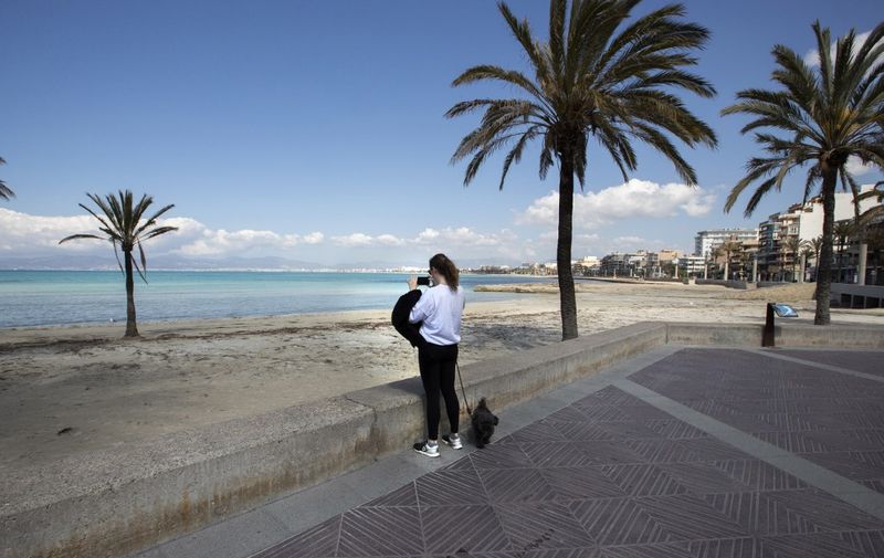 A woman walking a dog takes a picture of the empty Palma Beach, in Palma de Mallorca, on April 4, 2020, during a national lockdown to prevent the spread of the COVID-19 disease. - Spanish Prime Minister Pedro Sanchez announced today the extension of the country's lockdown until April 25 in order to curb the spread of the novel coronavirus. However, Spain today recorded a second successive daily fall in coronavirus-related deaths with 809 fatalities. The total number of fatalities  in the country stands at 11,744, second only to Italy. The number of new Spanish cases also slowed to 7,026, taking the total to 124,736. (Photo by JAIME REINA / AFP)