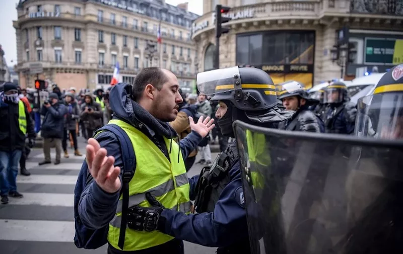 A protester wearing a yellow vests (gilets jaunes) gestures as a French Police officer conducts a search during a demonstration against rising costs of living they blame on high taxes, at the place de l'Opera, in Paris, on December 15, 2018. - The "Yellow Vests" (Gilets Jaunes) movement in France originally started as a protest about planned fuel hikes but has morphed into a mass protest against President's policies and top-down style of governing. (Photo by Lucas BARIOULET / AFP)