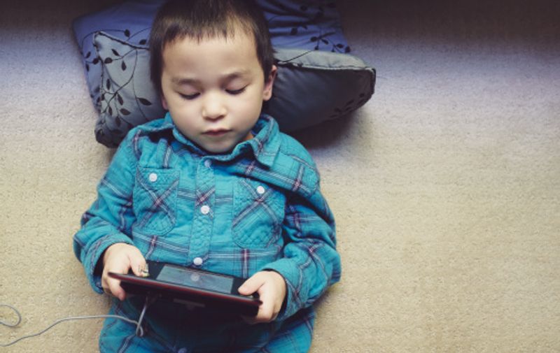 Child laying on carpet and pillow playing plugged in handheld gaming computer device.