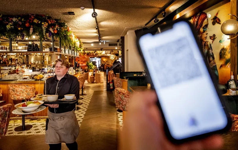 A customer holds a smartphone with a sanitary or health pass visible as a waiter walks past in a restaurant in The Hague on September 25, 2021, as some coronavirus (Covid-19) measures are lifted. - Keeping a distance of 1,5 meters is no longer mandatory, the catering industry can open again with maximum capacity and people must show a sanitary or health pass when visiting resteraunts, cafes and events. (Photo by Koen van Weel / ANP / AFP) / Netherlands OUT