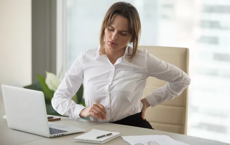 Upset young businesswoman feeling backache touching aching back muscles suffering from low-back lumbar pain sitting in incorrect posture on uncomfortable office chair, sedentary work problems concept
