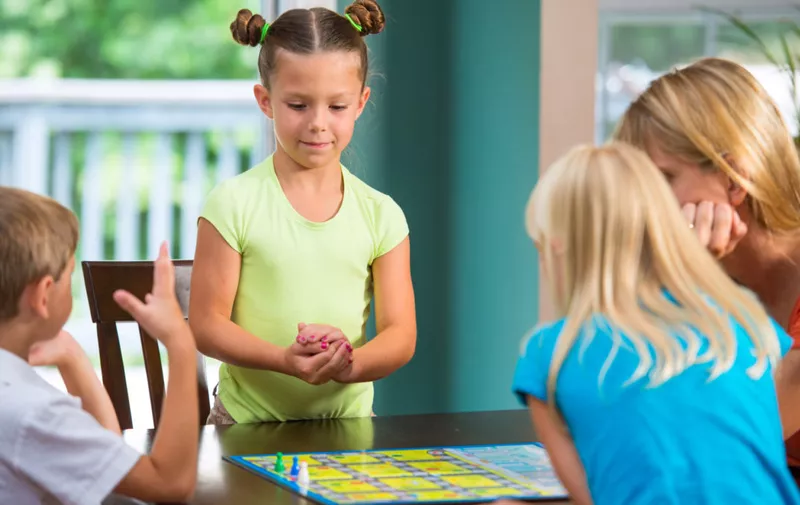 Authentic, fun image of a family playing a board game in a typical american home. Little girl with buns in hair shakes dice in her hands before her roll.