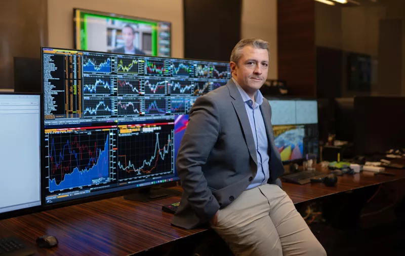 Poslovni čovjek u sivom sakou sjedi kraj stola s računalnim zaslonima koji prikazuju burze i financijske grafikone.