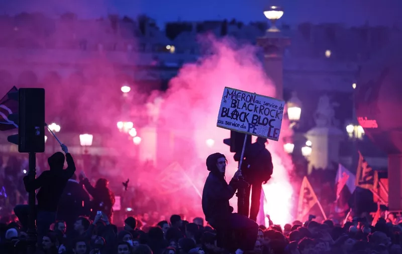 A protester on a traffic light holds a placard reading "Macron at the service of Black Rock, Black bloc at the service of the people" during a demonstration on Place de la Concorde after the French government pushed a pensions reform through parliament without a vote, using the article 49,3 of the constitution, in Paris on March 16, 2023. - The French president on March 16 rammed a controversial pension reform through parliament without a vote, deploying a rarely used constitutional power that risks inflaming protests. The move was an admission that his government lacked a majority in the National Assembly to pass the legislation to raise the retirement age from 62 to 64. (Photo by Thomas SAMSON / AFP)