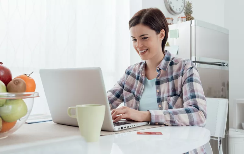 Smiling young woman connecting with a laptop in the kitchen at home, she is doing online shopping and buying products using a credit card